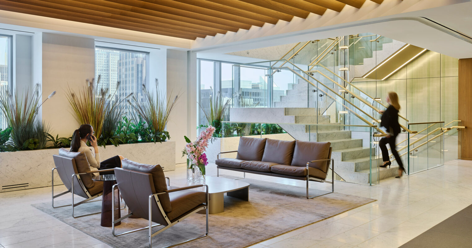 A modern office lobby featuring a recessed ceiling with wooden slats, marble stairs with glass railings, and seating areas with cityscape views.