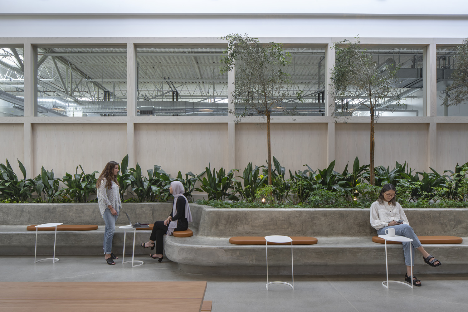 Spacious lobby with a biophilic design featuring an indoor garden bed, wooden paneling, and a skylight. Modern benches and side tables accommodate visitors amid the lush greenery, combining functionality with a connection to nature.