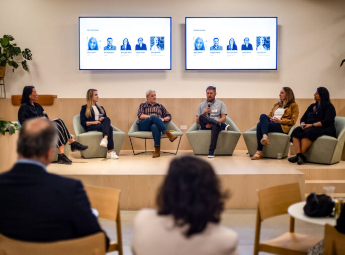 Six individuals lead a panel discussion at the Belkin El Segundo workplace. The interior space is brightly lit with warm wood accents and biophilic elements like robust plantings.