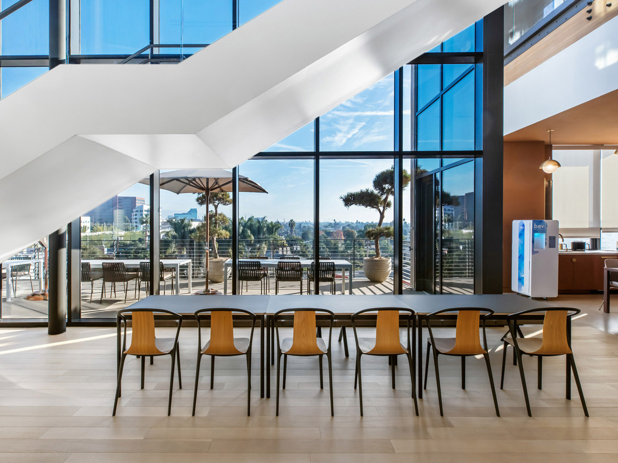 Sleek, modern interior with a floating white staircase, hardwood floors, and expansive windows offering abundant natural light and exterior views. Mustard-yellow chairs surround a long communal table, emphasizing a contemporary, collaborative space.
