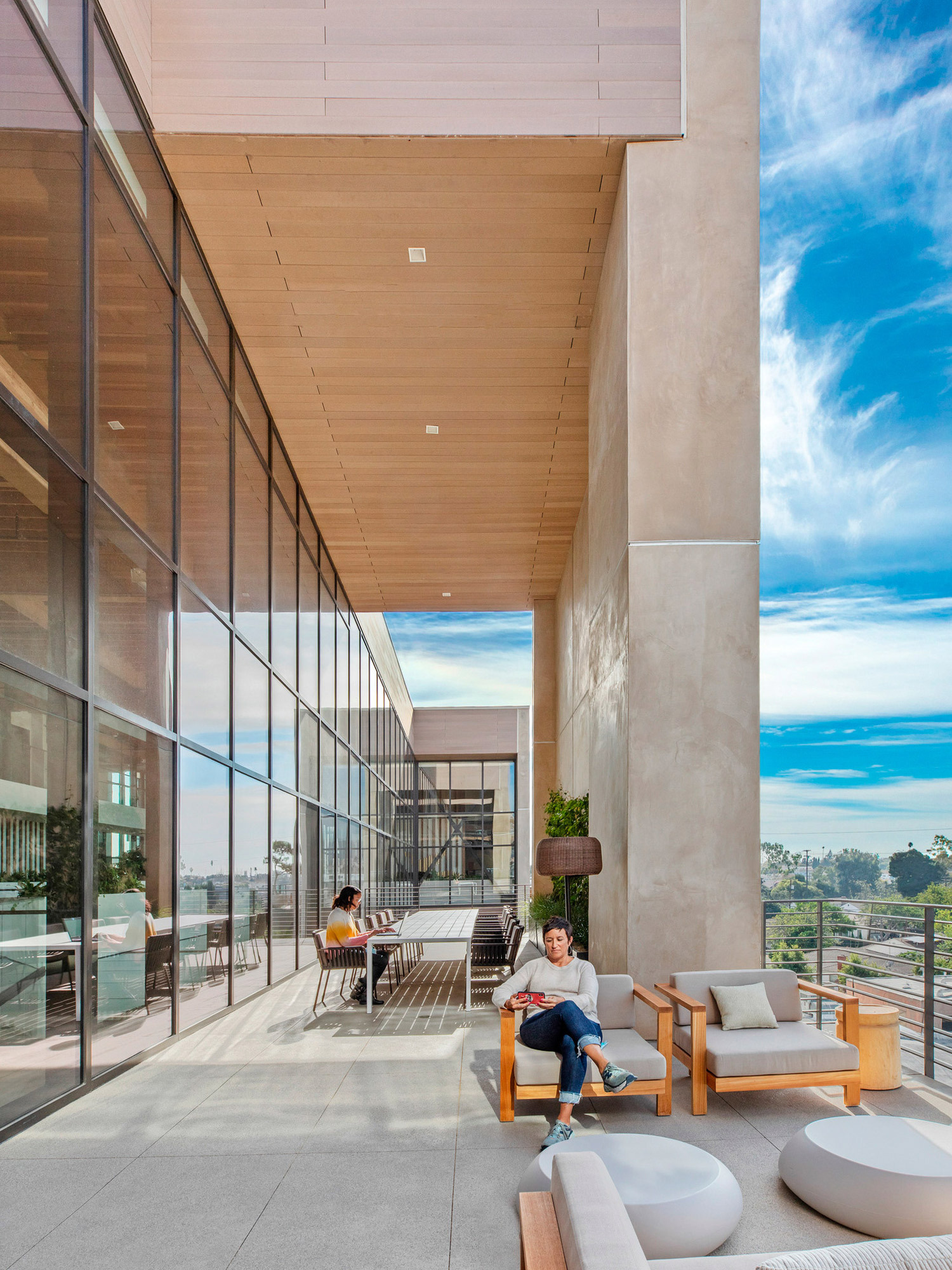 Spacious outdoor terrace with a minimalist design, featuring sleek beige and white furniture. Wooden panels line the ceiling, complementing the beige pillars and floor-to-ceiling windows, which offer a view of clear skies and surrounding greenery.