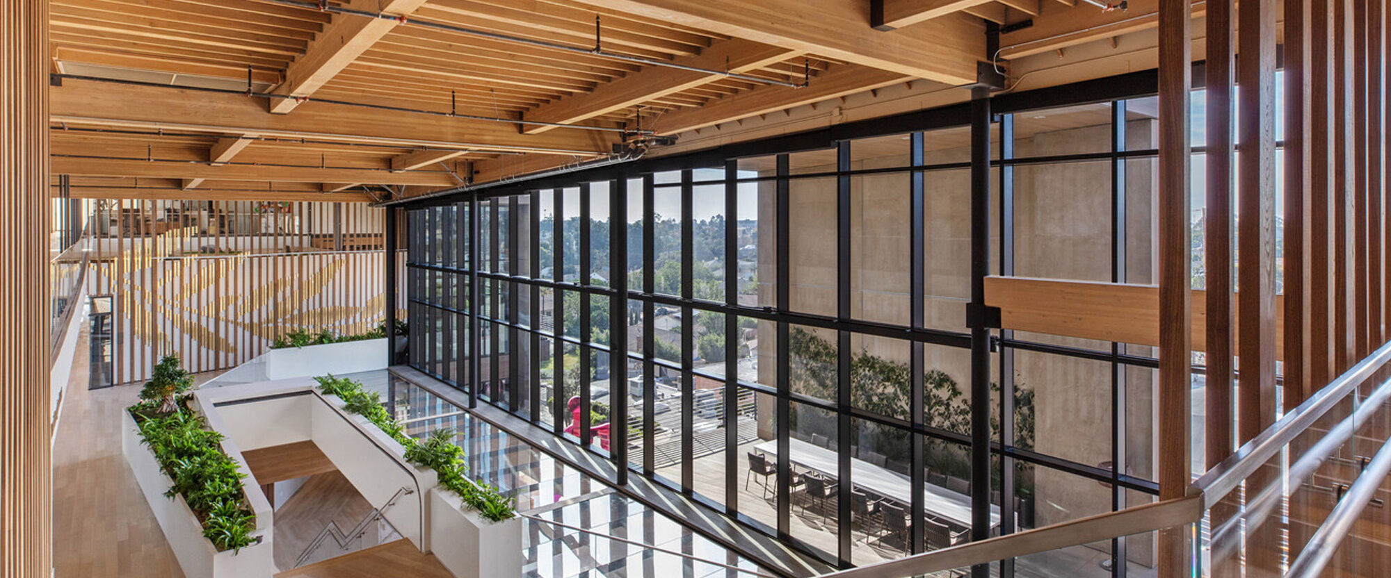 Modern office interior highlighting exposed wooden beams and slatted ceiling design, complemented by glass railing, open-plan staircases, and indoor greenery, creating a biophilic, industrial aesthetic with natural light.
