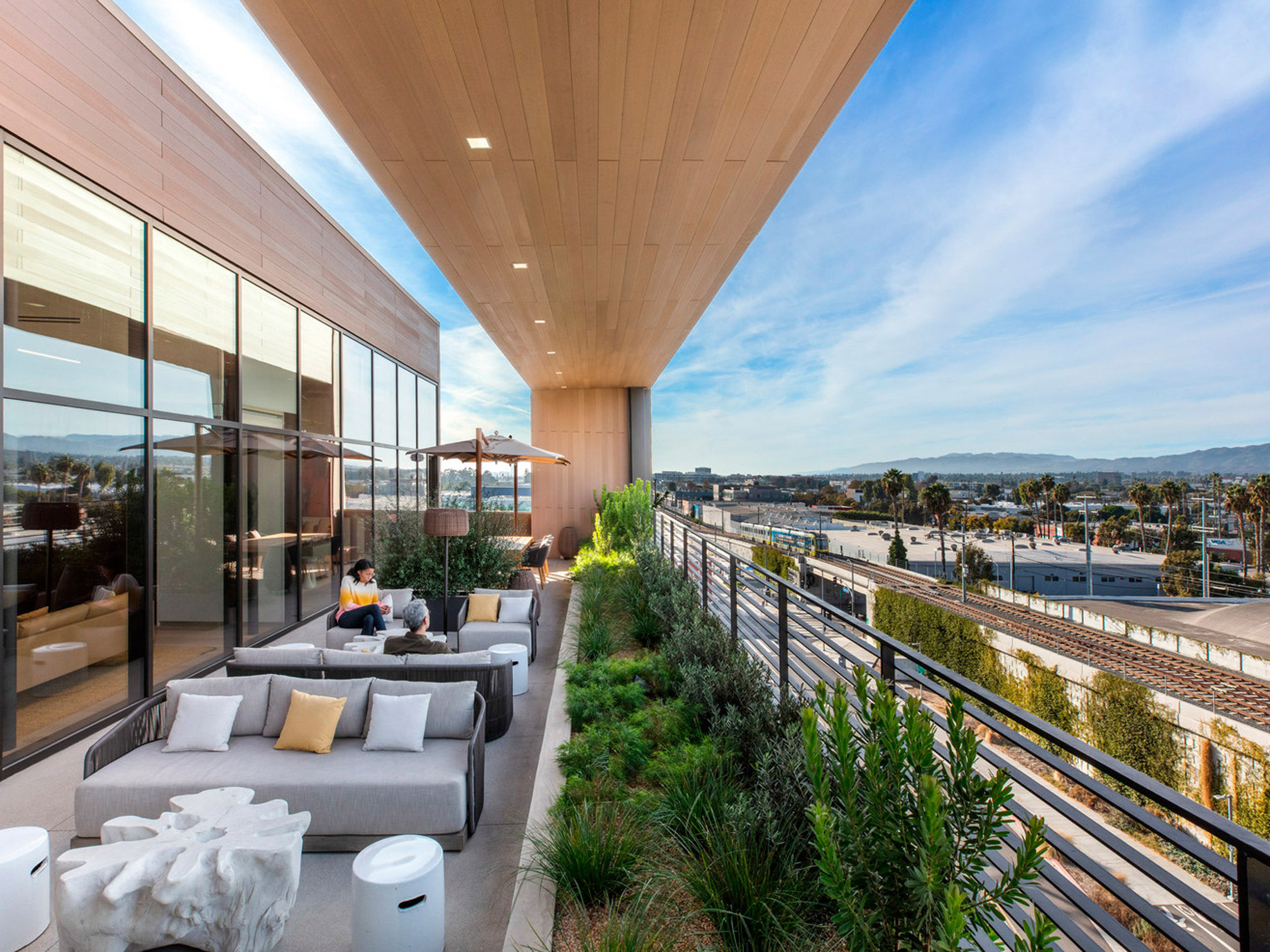 Open-plan terrace featuring floor-to-ceiling glass walls, minimalist furniture with neutral upholstery, and wooden ceiling extending outdoors, harmonizing with panoramic views of the urban skyline. Well-positioned greenery adds a touch of nature to the modern space.
