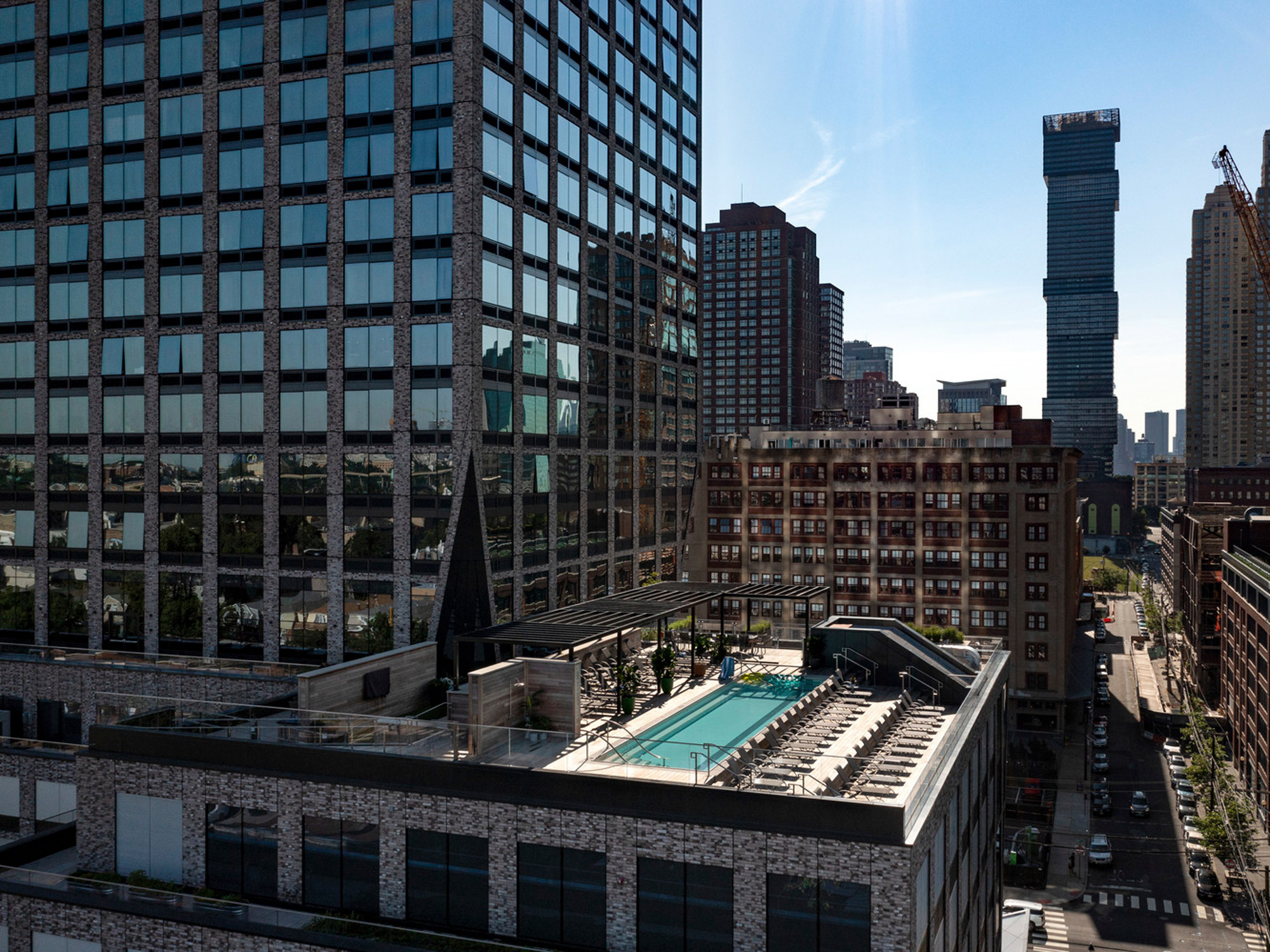 Rooftop terrace with a rectangular swimming pool reflecting the clear sky, flanked by lounge chairs, amidst a backdrop of mixed architectural styles from neighboring high-rise buildings, highlighting urban design integration.