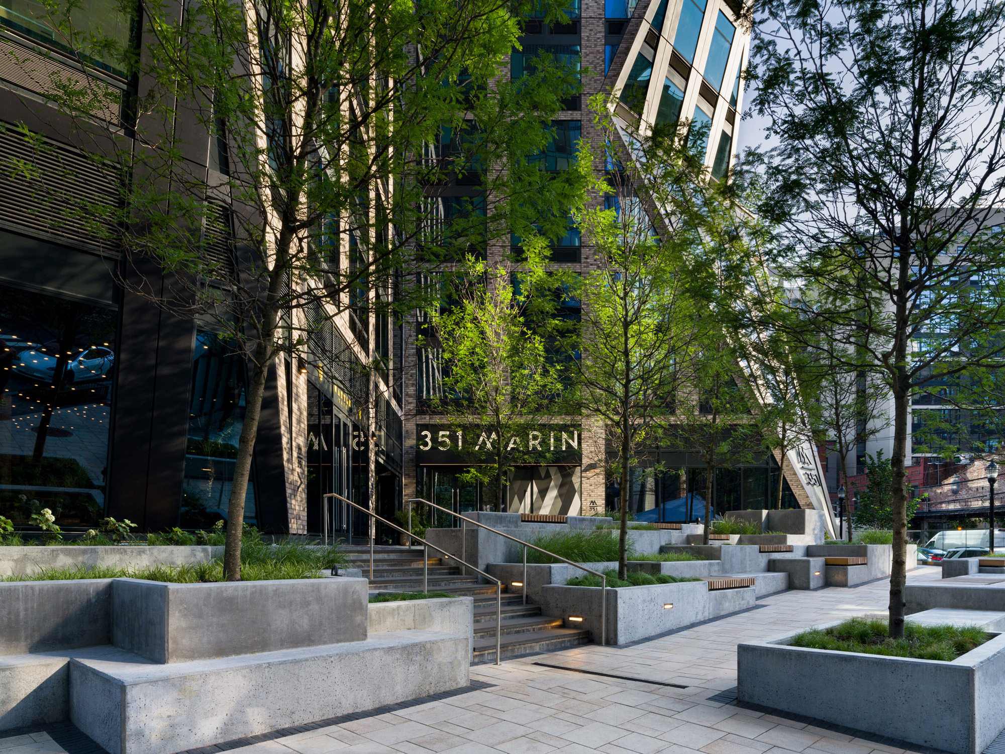Urban landscape design featuring a series of concrete planters and integrated seating, enhancing the pedestrian experience at the entrance of a modern building. Young trees and assorted greenery soften the hardscape, adding a touch of nature to the setting. The architecture displays a mix of reflective glass and metal elements, with building signage prominently displayed. The area is complemented by a stone pathway and subtle lighting, inviting passersby to a serene enclave amidst the city bustle.