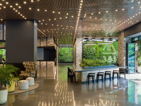 Contemporary lounge area boasting an illuminated pegboard ceiling and glossy floor, with a green living wall featuring numerical art, flanked by industrial-style seating and vibrant planters.