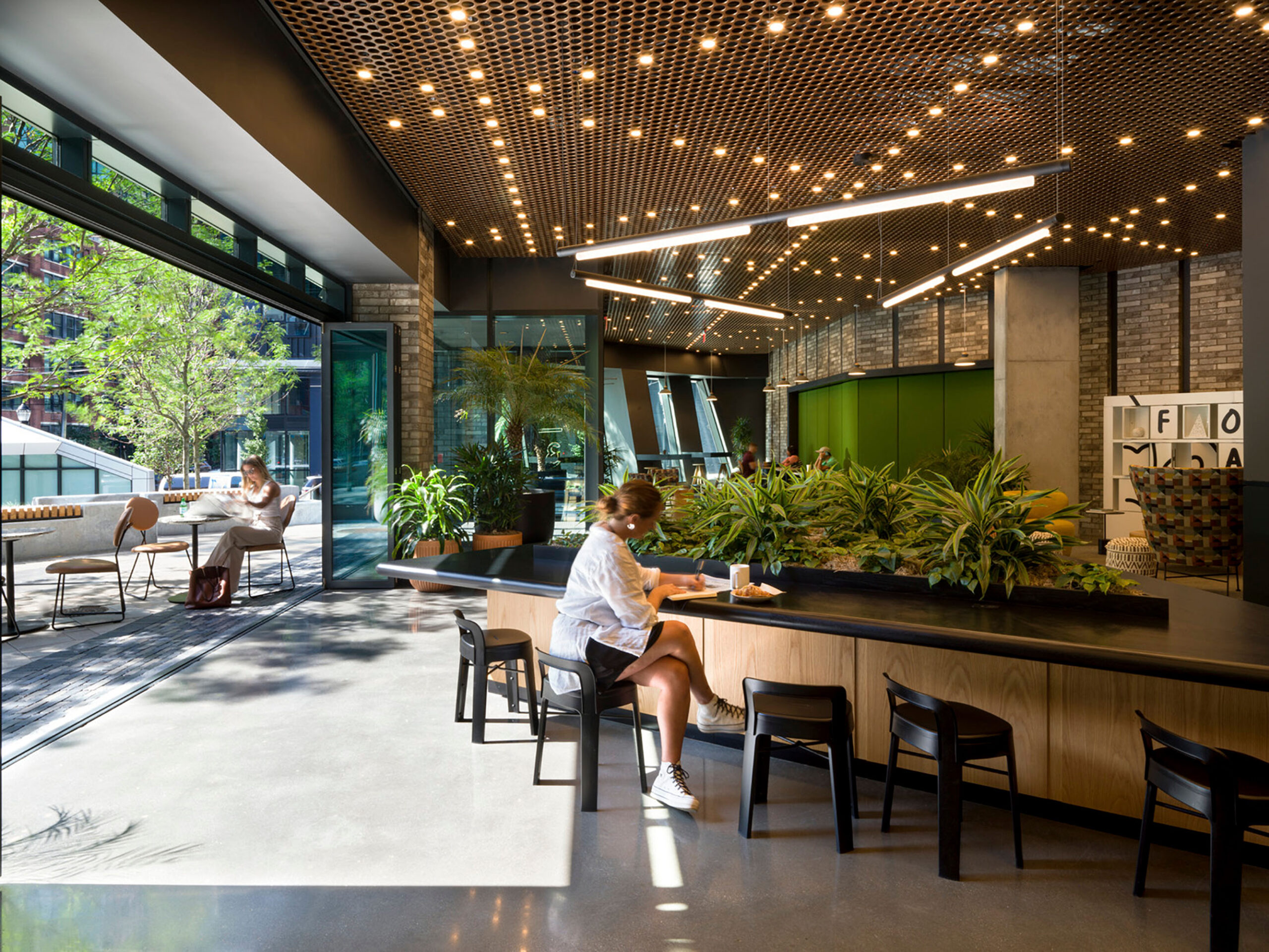 A modern and inviting indoor space illuminated by natural light and intricate ceiling lighting. The room has open access to the outside and is filled with plants and biophilic elements.