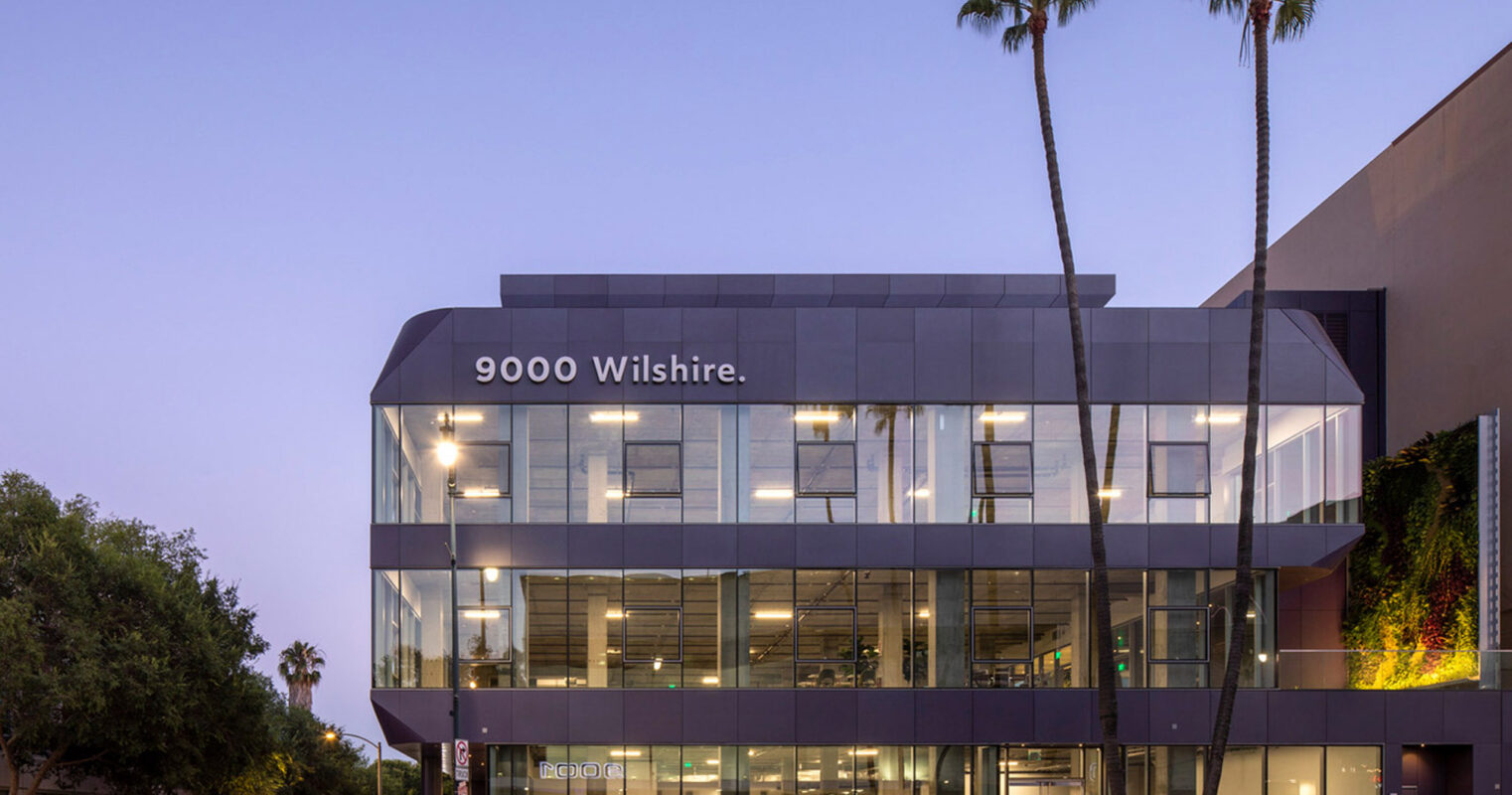 Modern three-story commercial building at dusk with extensive use of glass and geometric lines, illuminated interior showcasing a sleek, minimalist design, flanked by tall palm trees against a twilight sky. Address "9000 Wilshire" prominently displayed.