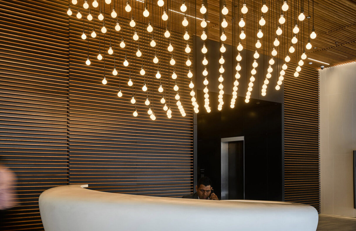 Modern reception area featuring a sleek, curved white desk against a backdrop of vertical wooden slats. Overhead, a striking array of suspended cylindrical lights forms a dynamic, cascading chandelier, enhancing the space with warmth and a contemporary aesthetic.
