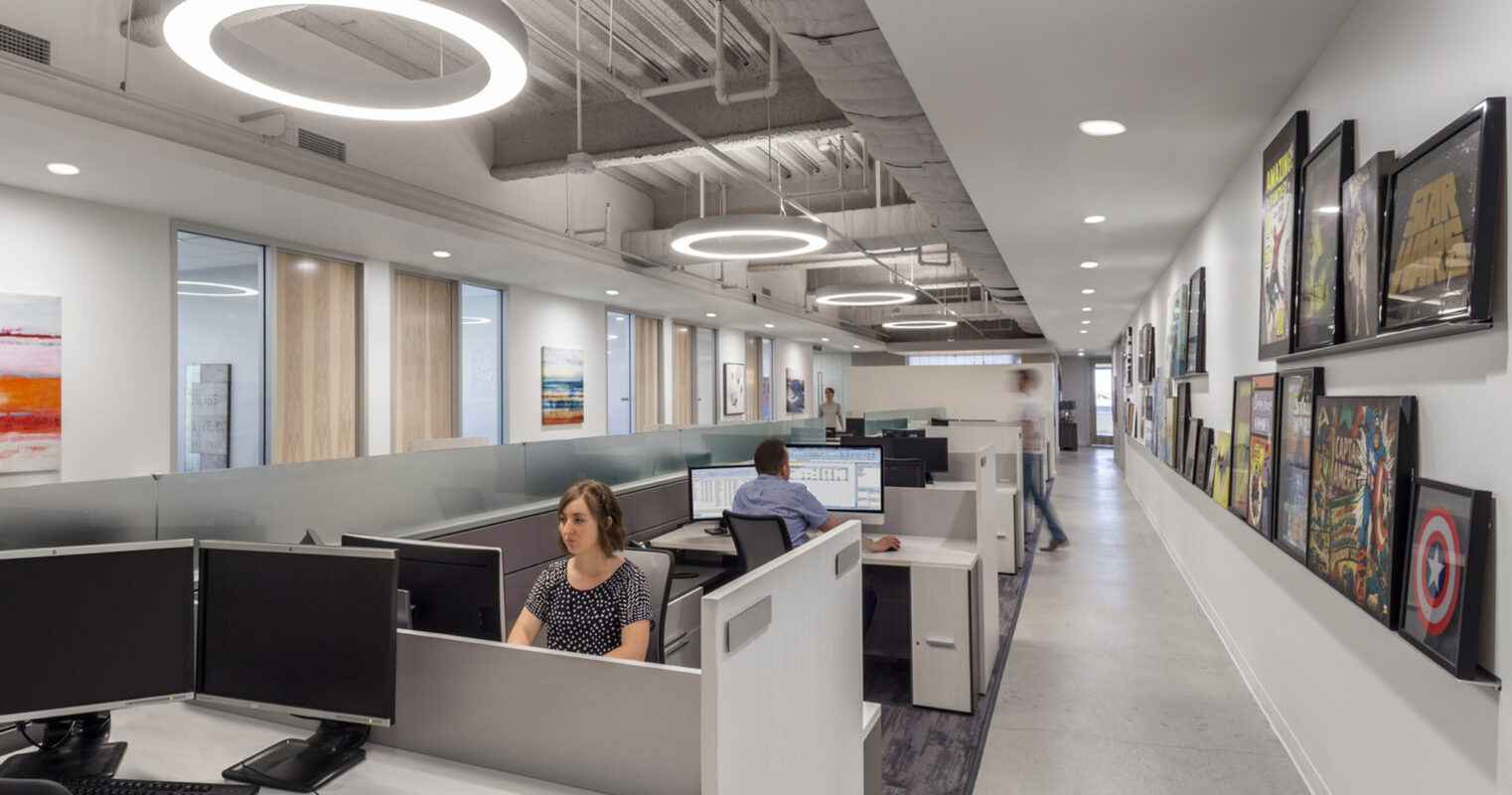 Modern open-plan office featuring streamlined workstations with ergonomic chairs, circular overhead lighting, and an exposed ceiling, complemented by a gallery wall of vibrant artwork enhancing the creative ambience.
