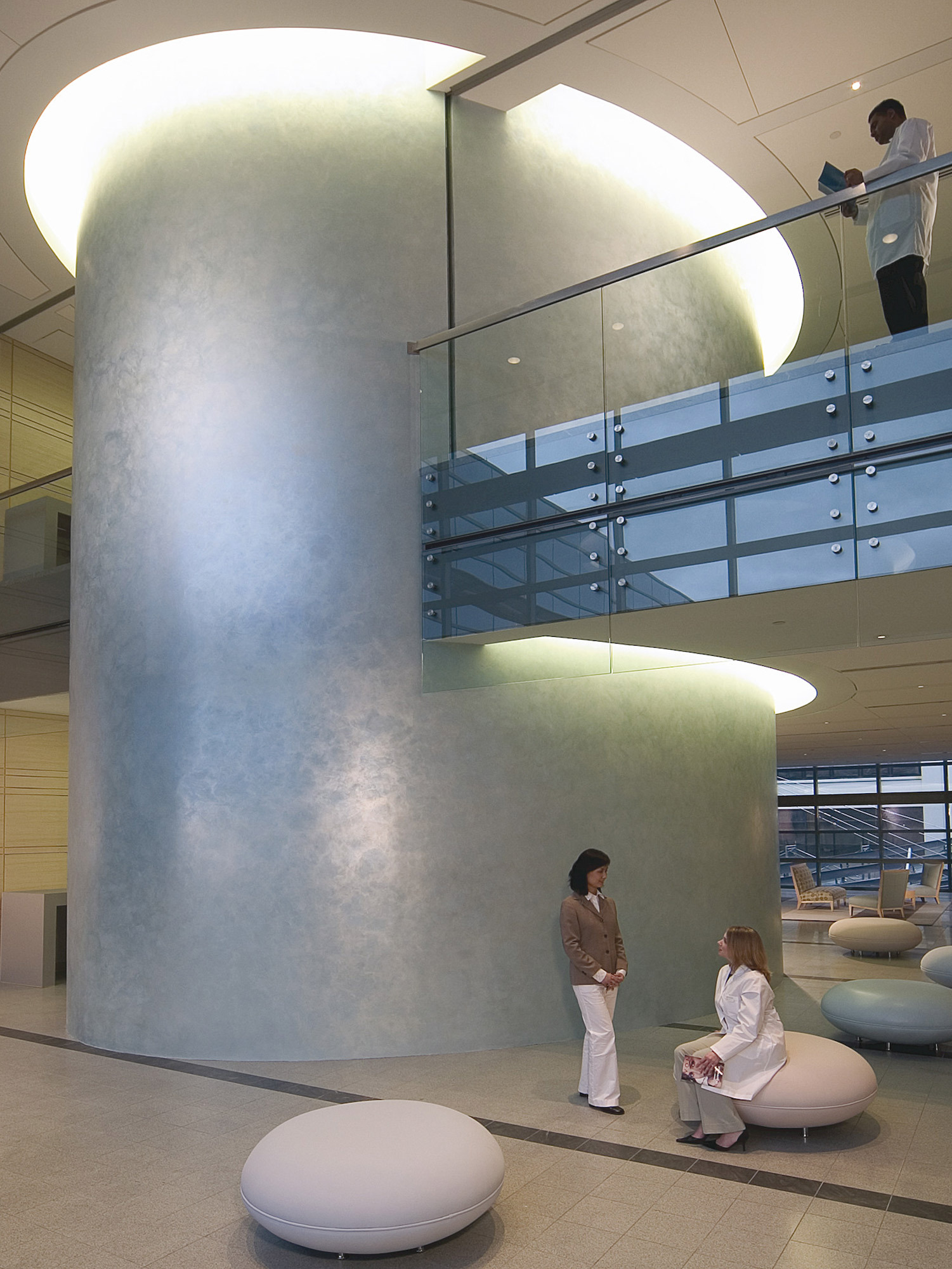 Modern lobby area with a cylindrical central feature, subtle lighting creates depth, enhancing the frosted finish. Sleek chrome stair rails complement the minimalist style while the use of white oval seating adds a contemporary touch to the space.