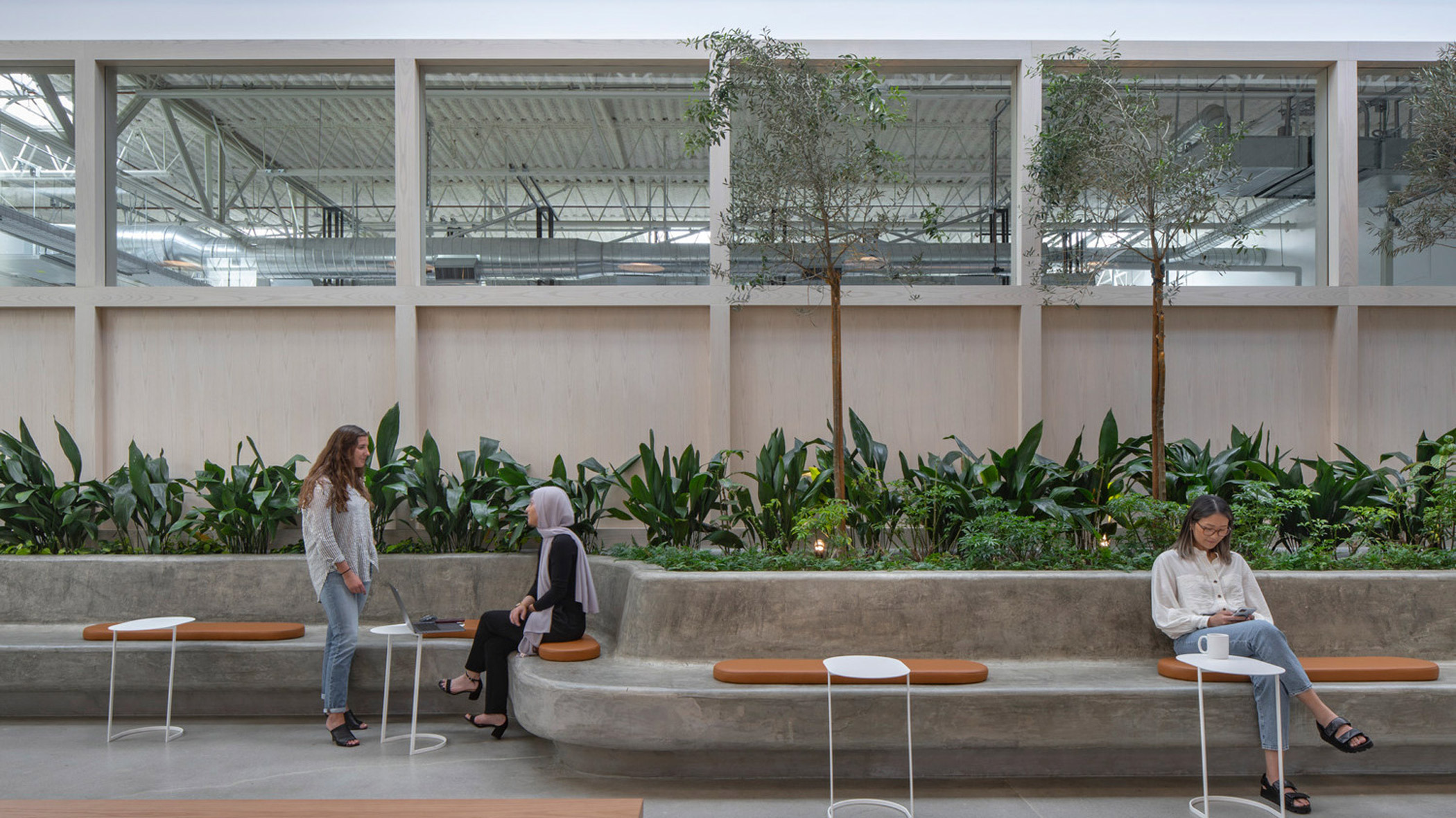 Spacious lobby with a biophilic design featuring an indoor garden bed, wooden paneling, and a skylight. Modern benches and side tables accommodate visitors amid the lush greenery, combining functionality with a connection to nature.