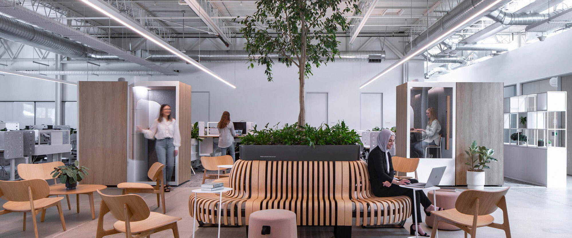 Spacious office interior bathed in natural light, featuring an eclectic mix of modern wooden desks, upholstered couches, and an indoor planting area offering a blend of functionality and biophilic design. Overhead exposed ductwork complements the industrial-chic aesthetic.