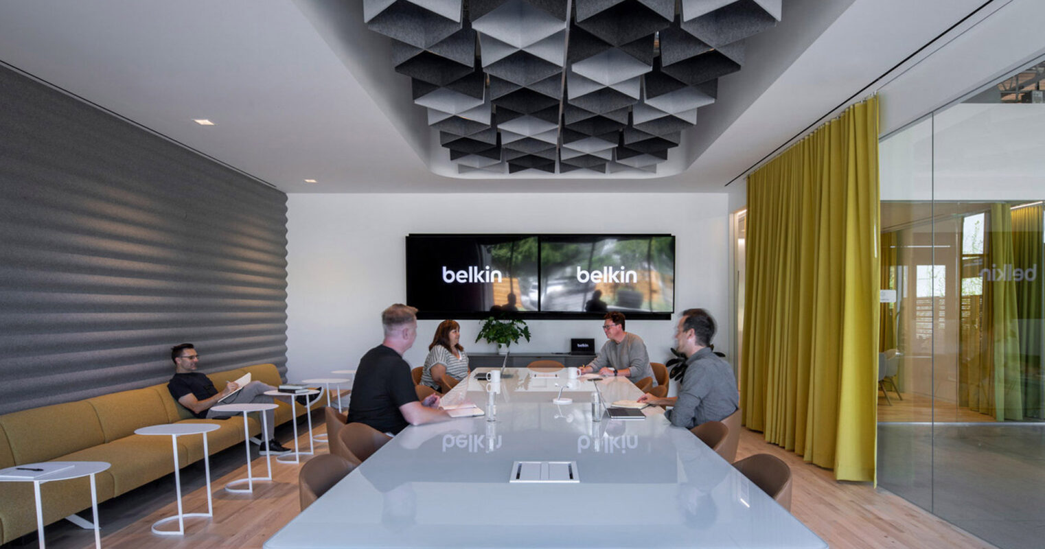 Modern corporate meeting room featuring a sleek wooden table, angular black acoustic ceiling panels, a digital screen for presentations, and full-height glass walls. Neutral tones are complemented by pops of color from mustard draperies and upholstered office chairs.