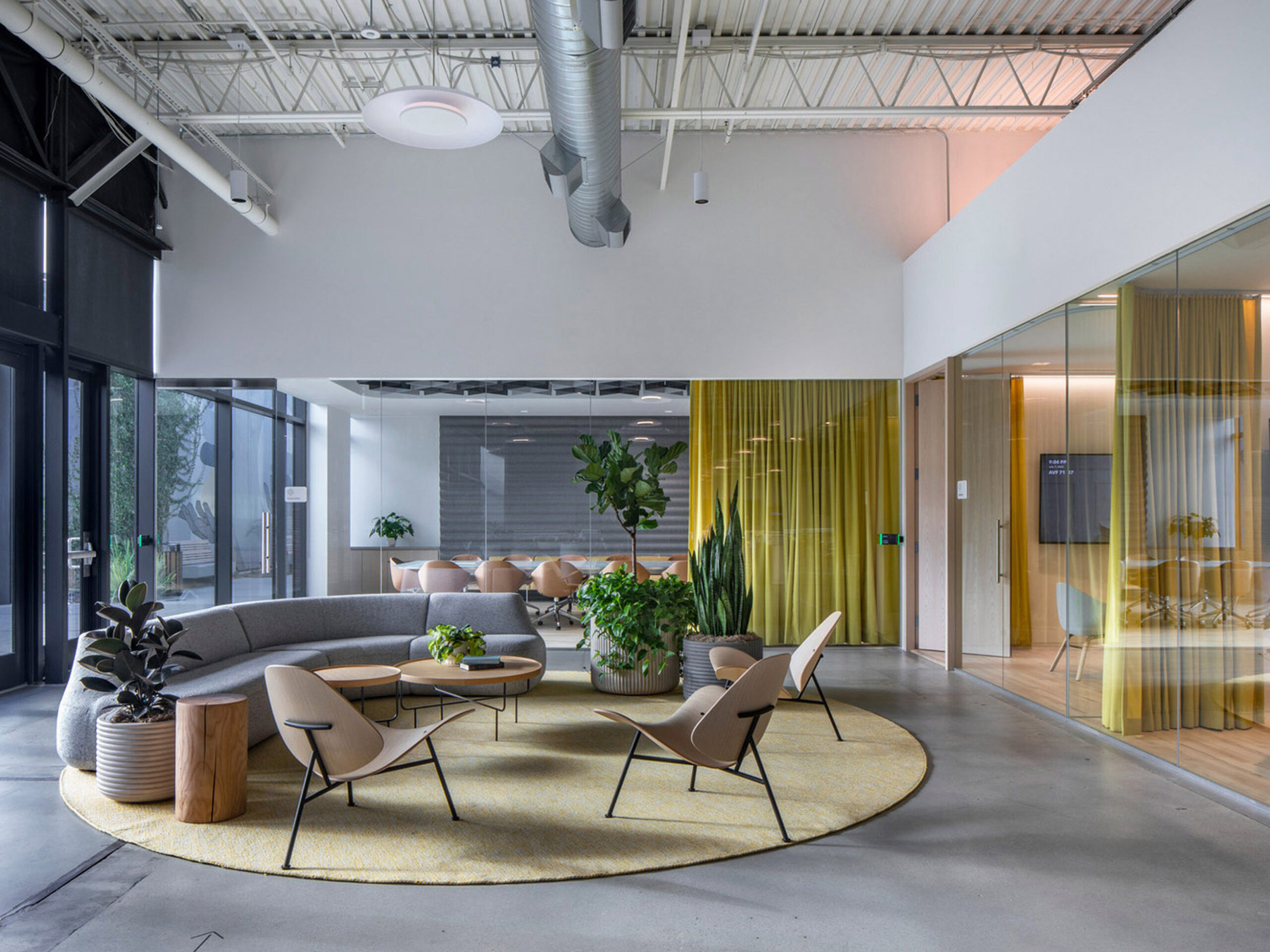 Modern open-plan office lounge with a curved gray sofa and tan armchairs centered around a circular yellow rug. Industrial ceiling, vibrant green office dividers, and potted plants add a touch of nature to the sleek environment.