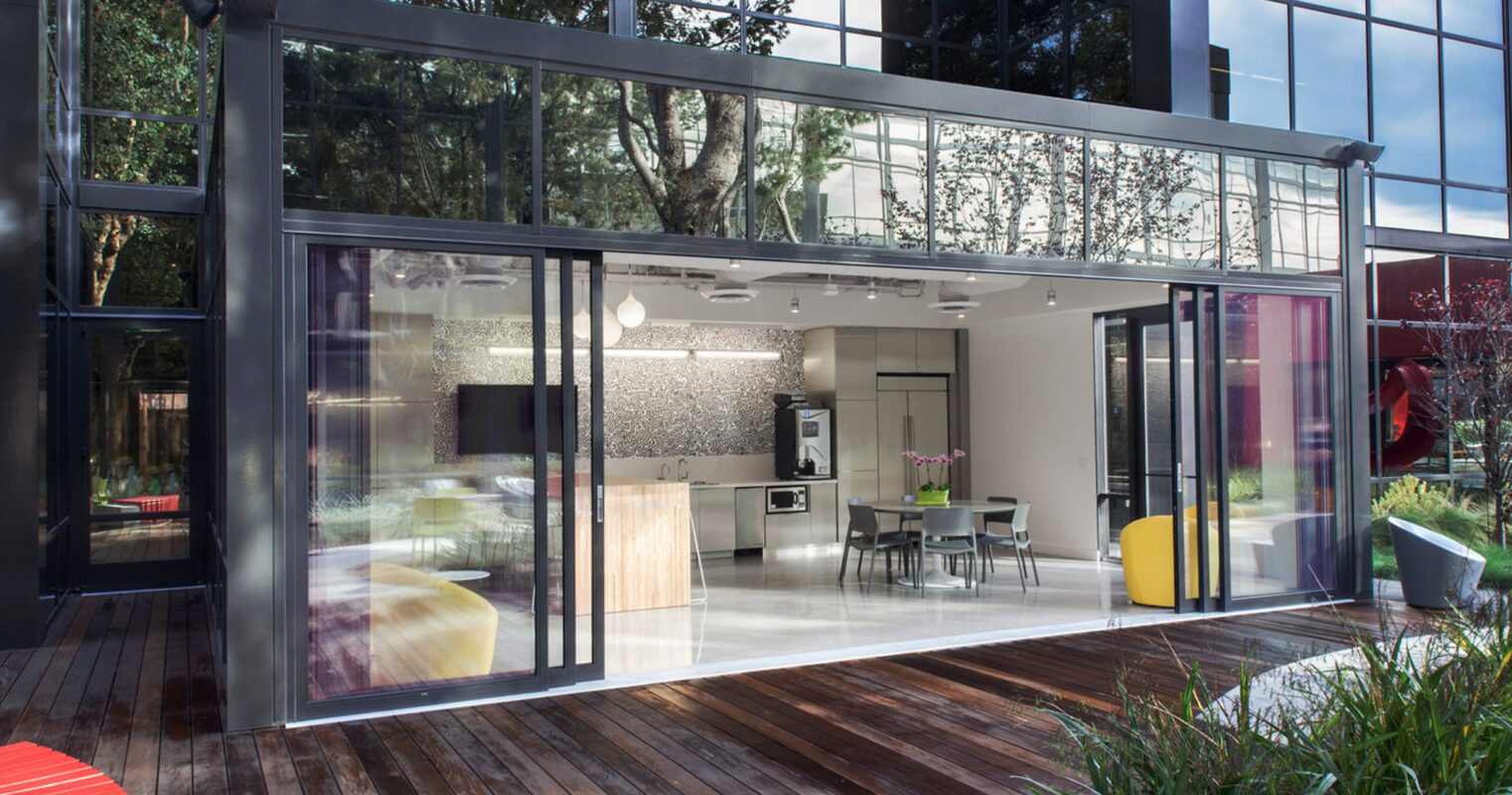 Modern open-concept living space with retractable glass doors blending indoor and outdoor areas. Sleek kitchen visible in background, complemented by contemporary furniture and hardwood flooring throughout. Vibrant accent seating adds a pop of color to the natural palette.