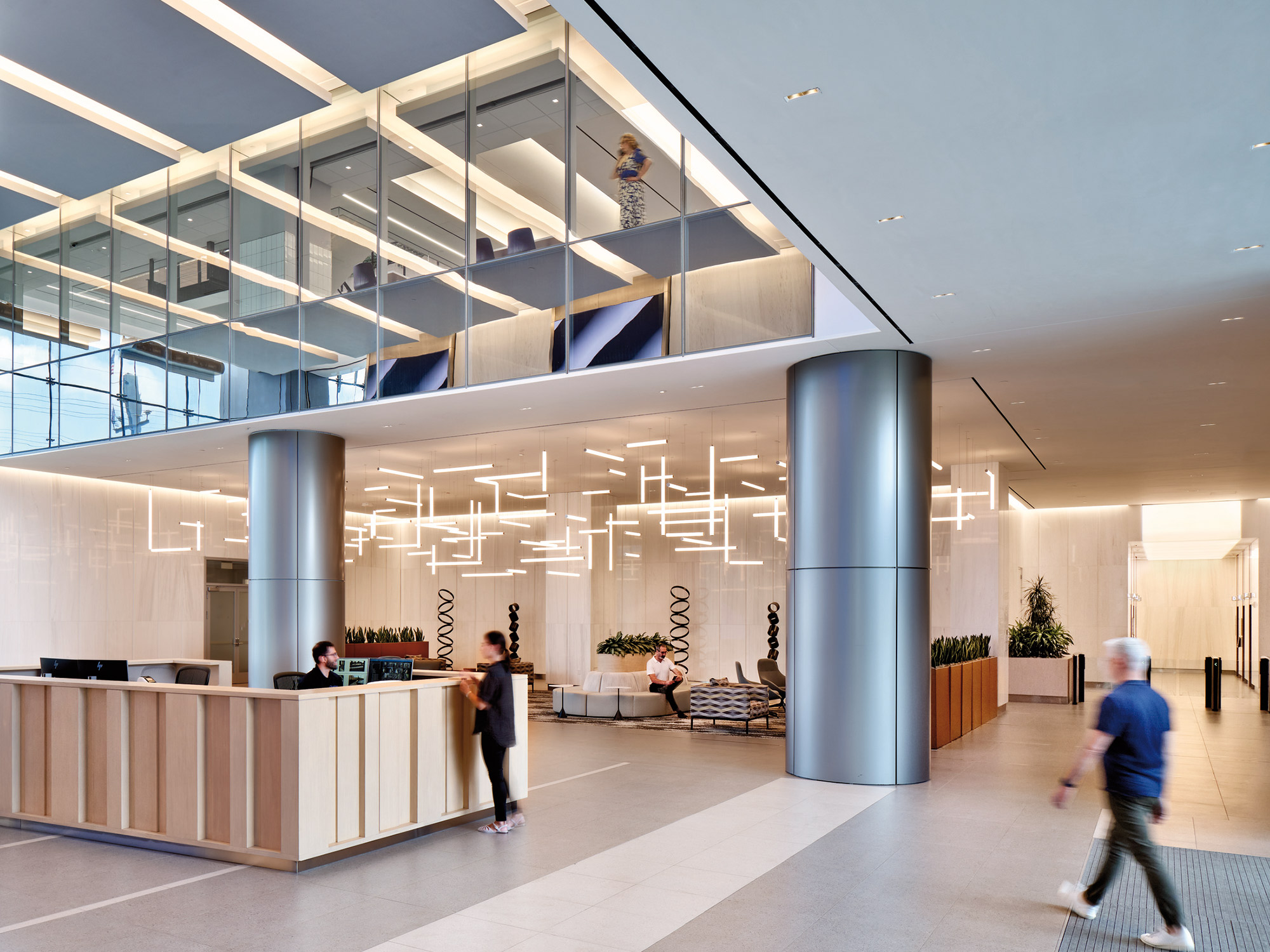 A sleek and modern corporate lobby featuring a reception desk, stylish hanging lights, and multi-level walkways with glass balustrades, with people entering and moving about the space.