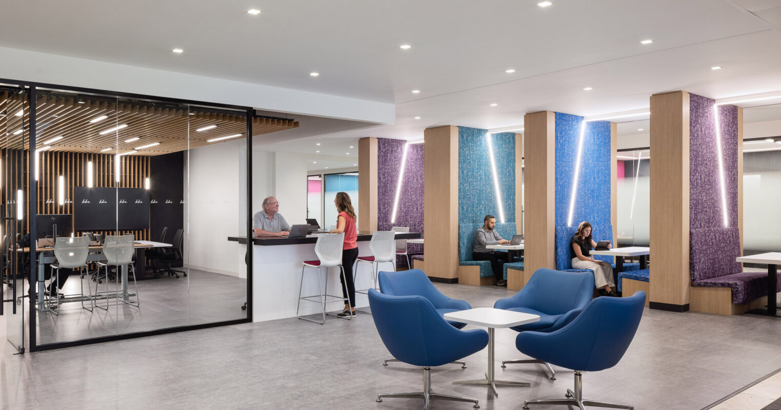 Contemporary office break room with vibrant blue egg chairs, sleek white high-top tables, and people in casual conversation. Textured columns with purple accents complement the neutral-toned flooring and wooden slatted ceiling design.