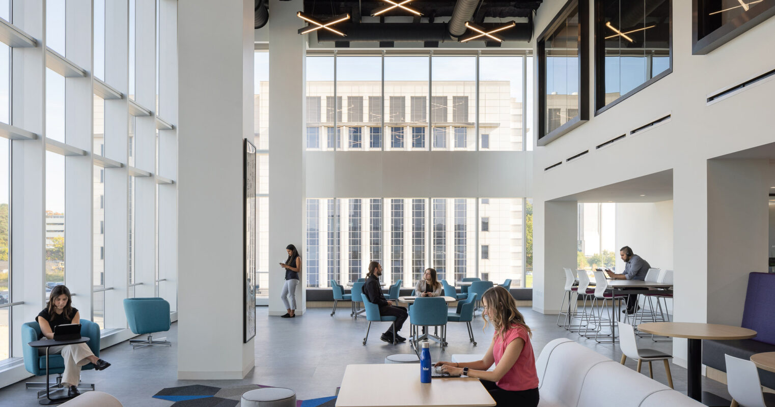 Bright, modern office lounge with geometric pendant lighting, floor-to-ceiling windows, and a mix of casual seating arrangements featuring sofas and cushioned chairs. Varied carpet patterns add pops of color, enhancing the space's energetic yet professional ambiance.