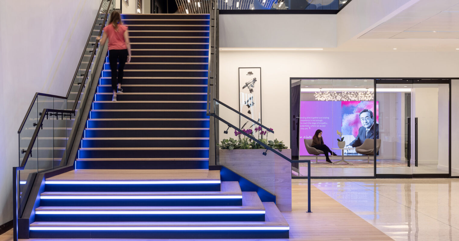 Modern office lobby featuring illuminated blue stair risers, creating a focal point. The stairs lead to a bright, open mezzanine with people casually interacting by a digital display wall, reflecting a dynamic and technologically-integrated workspace.