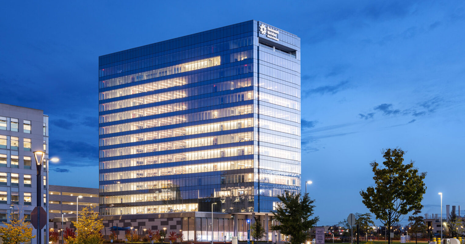 Twilight view of a modern glass-walled office building, showcasing reflective surfaces with interior lights creating a glowing effect, set against a deep blue sky. Landscaping and urban setting frame the architecture.