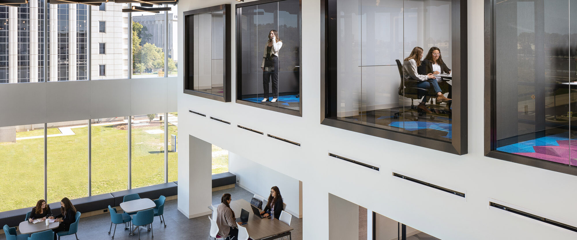 Modern office space featuring elevated, glass-walled meeting rooms with open-plan seating below. Exposed black ceiling infrastructure contrasts with white walls. Pops of color from scattered furniture accentuate the room's contemporary design and collaborative atmosphere.