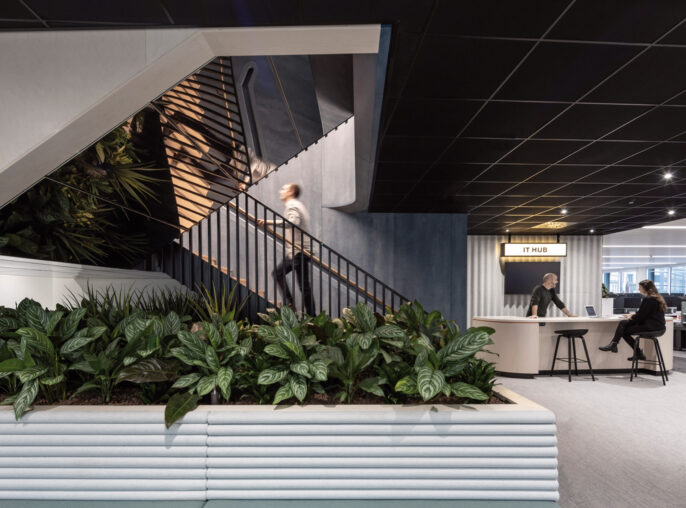 Modern office lobby with a sleek staircase featuring geometric railings, surrounded by lush greenery. The space evokes a dynamic work environment with its contrasting dark ceiling and soft blue upholstered seating area near the reception desk.