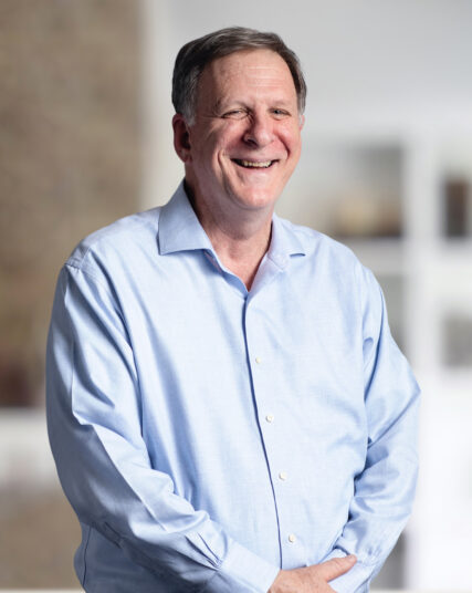 This photograph captures a man with a wide, sincere smile, wearing a light blue button-up shirt. He is standing with one hand in his pocket, against a backdrop that is light and indistinct, giving the impression of a relaxed yet professional setting.