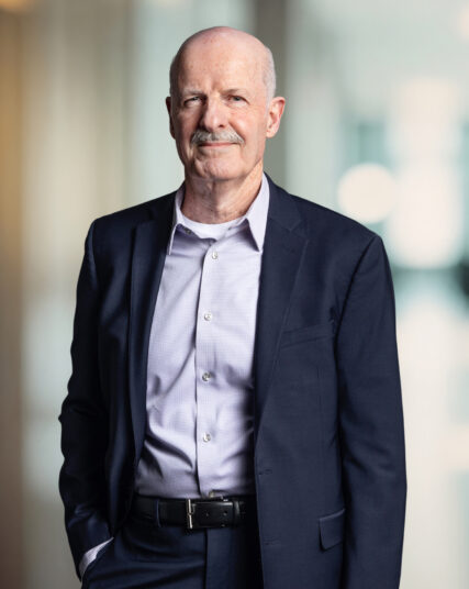 This image captures a distinguished looking man with a mustache, presenting a subtle smile and wearing a tailored dark suit with a light blue button-down shirt. He stands confidently, hands in pockets, against a background that is bright and blurred, likely an office setting with natural lighting.