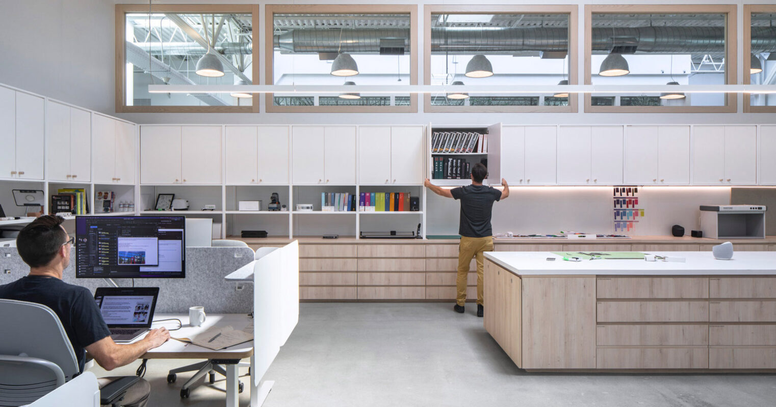Modern open-plan office featuring natural wood finishes, with workstations, shelving units, and pendant lighting. An individual interacts with material samples on the back wall, emphasizing the space's functional design intent.