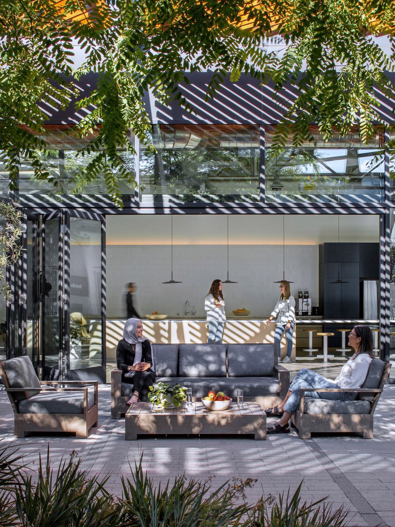 Modern outdoor lounge area with slatted wooden pergola casting shadows, featuring plush seating, glass walls, and a kitchenette, set amidst lush greenery for a seamless indoor-outdoor transition.