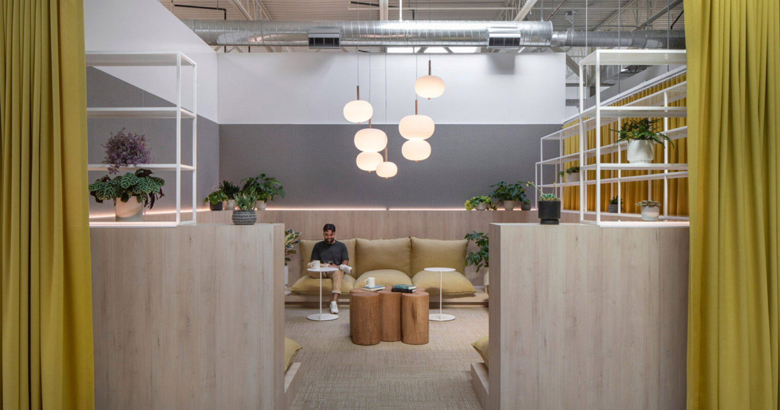Open-plan office space featuring minimalist wooden partitions, vibrant yellow drapery for privacy, and a comfortable seating area with a tan sofa. Overhead, spherical pendant lights provide soft illumination, complementing indoor greenery and enhancing the modern, tranquil atmosphere.
