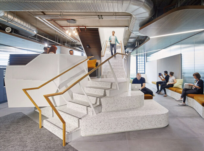 Modern office interior featuring a central floating staircase with white steps and gold accents. The surrounding open-concept space includes a lounge area with mustard yellow seating, exposed ductwork overhead, and people engaged in casual conversation, embodying a contemporary work environment.