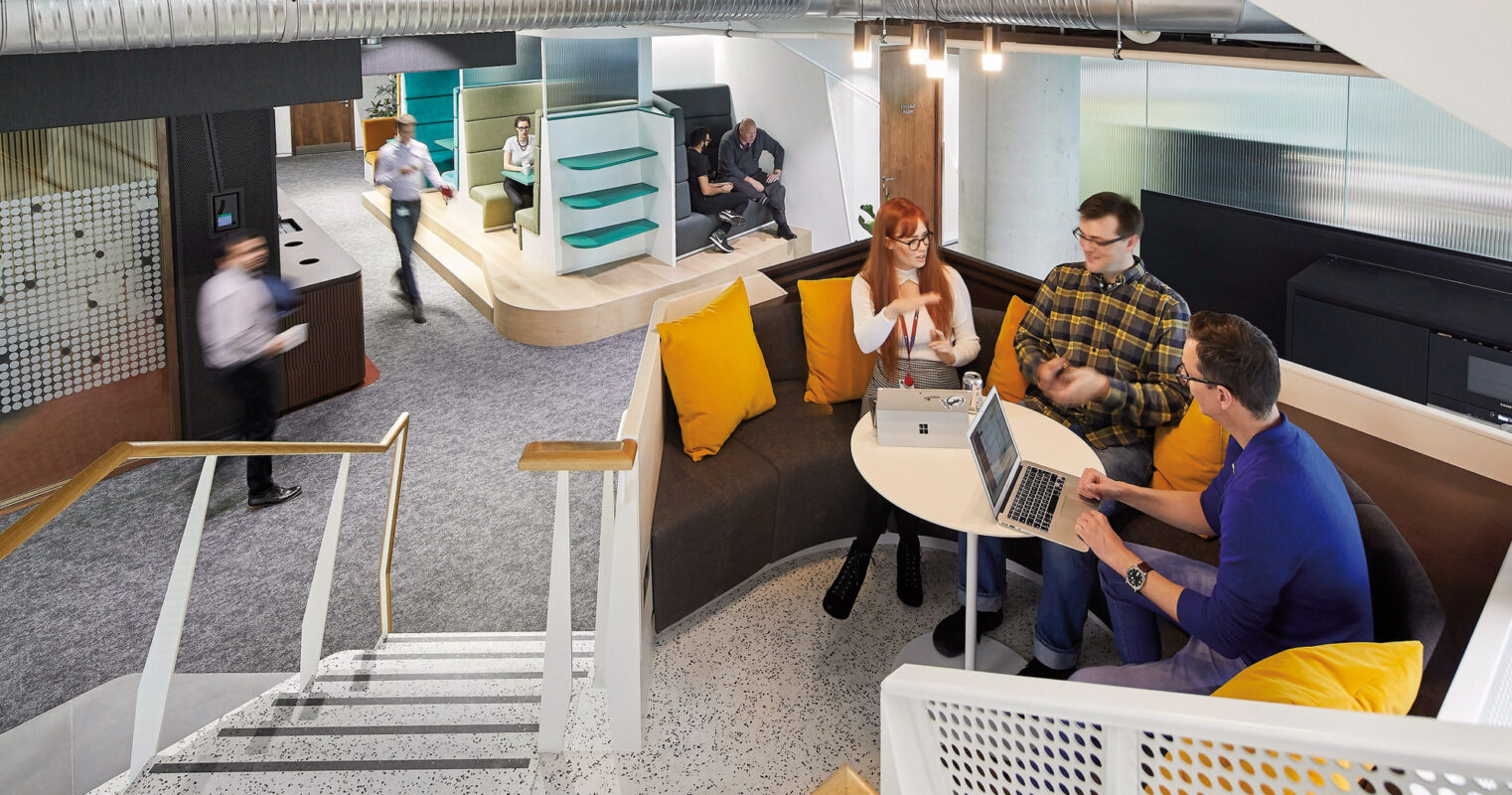 Modern office space with industrial aesthetic, featuring exposed ductwork and a speckled terrazzo floor. A central wooden table with yellow accents provides a collaborative work area, while a slide in the background adds playful functionality. Mesh room dividers offer privacy without visual obstruction.