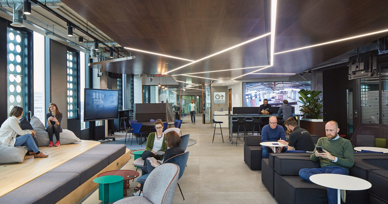 Modern office lounge area featuring tiered wooden seating, integrated LED lighting, and a mix of informal workspaces. The design encourages collaboration with circular seating accents and technological amenities, complemented by a palette of warm woods, greens, and neutral tones.