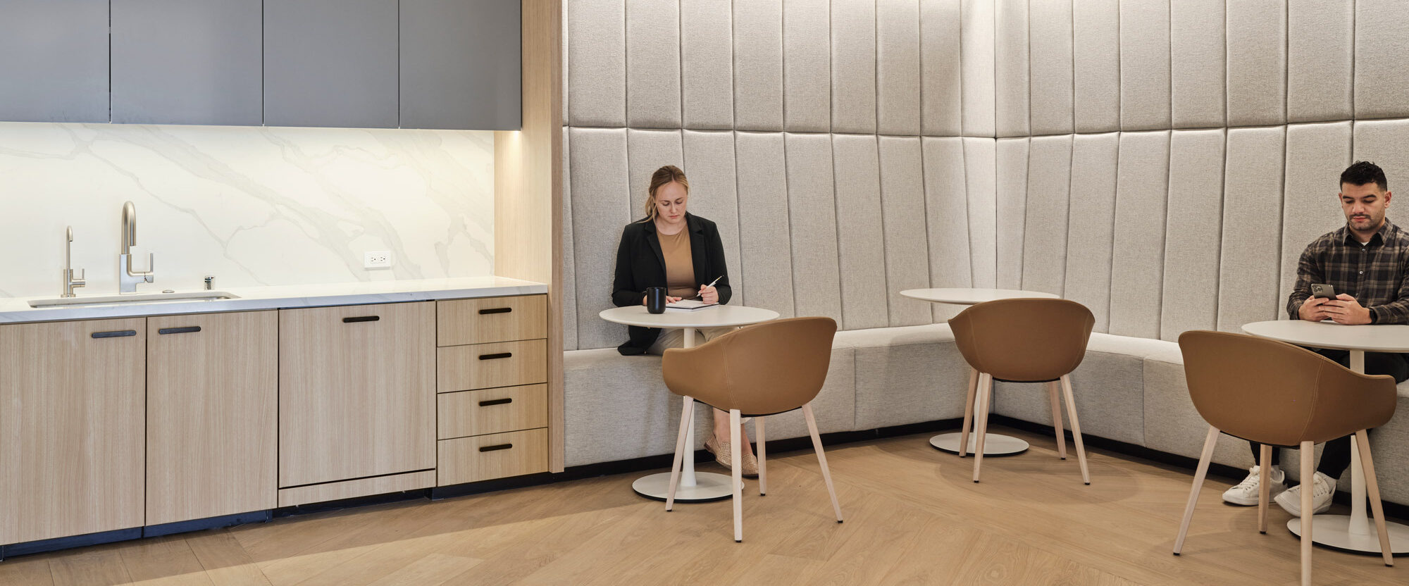 Modern kitchenette and dining area featuring sleek gray cabinetry, marble backsplash, and light wood tones. Padded wall panels add texture, while individuals quietly work at round tables, blending functionality with minimalist design.