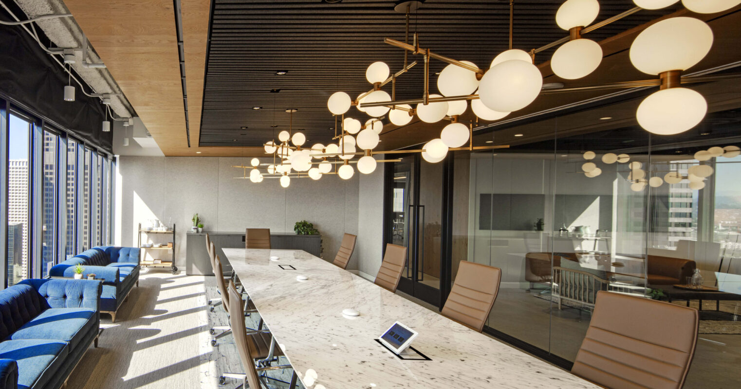 Modern conference room featuring a long, marble table with stylish orb-like pendant lights above. Sleek black chairs, wood ceiling panels, and floor-to-ceiling glass walls provide a panoramic city view, complemented by cozy blue seating along the perimeter.