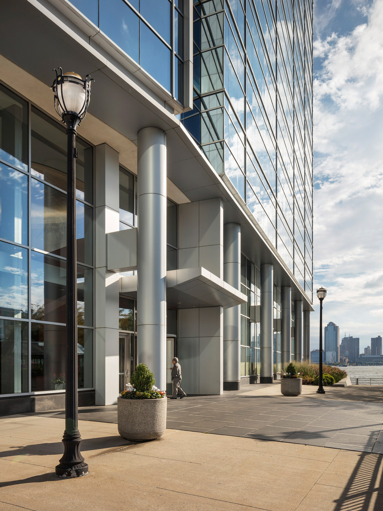 Modern commercial building entrance featuring reflective glass façade, cylindrical steel columns, and large planters. A person walks by, adding scale to the grand, minimalist architecture against a backdrop of clear blue skies.
