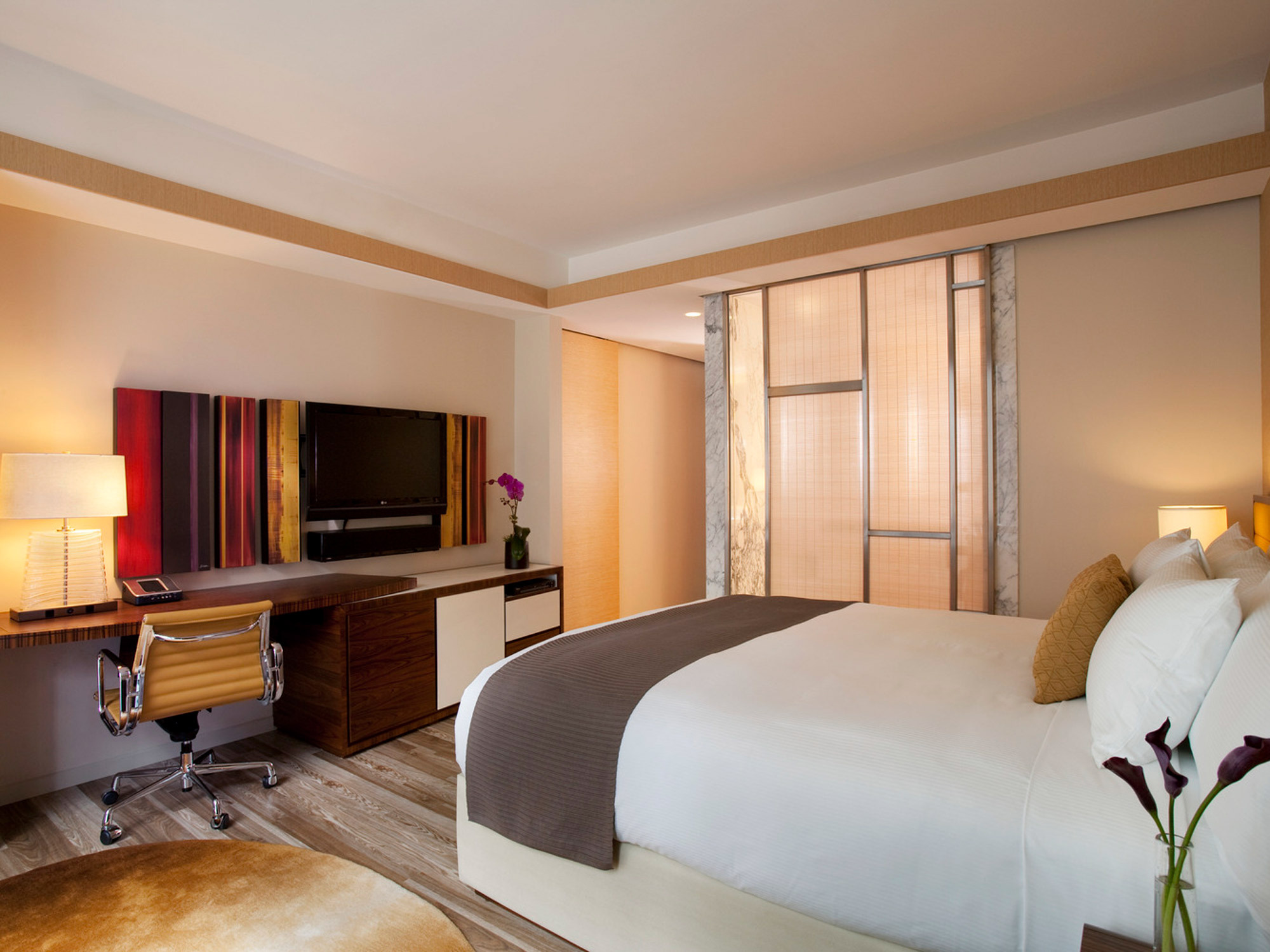 Warm-toned contemporary bedroom featuring a plush king-sized bed with beige and grey bedding, flanked by matching wood nightstands and soft-glow table lamps, opposite a sleek wooden entertainment console and a large flat-screen TV, with natural light filtering through translucent window shades.