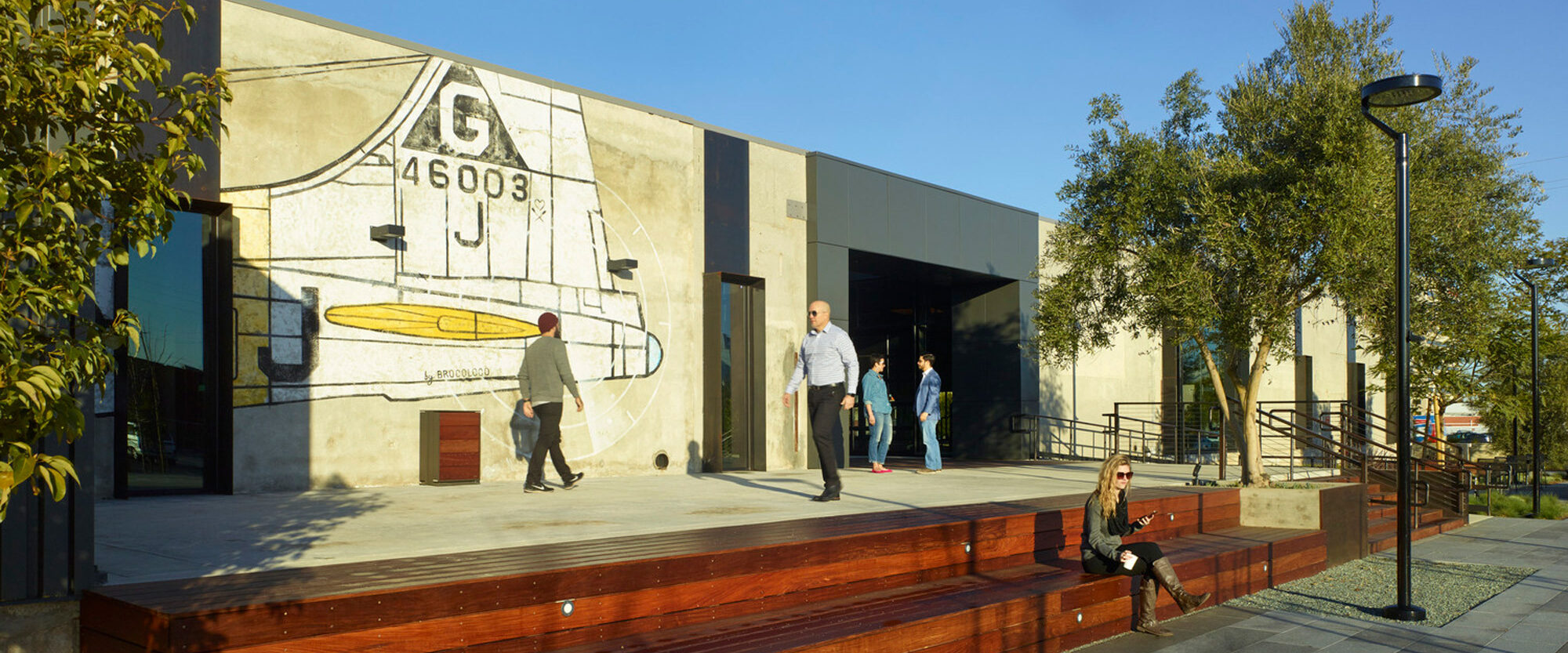 Modern urban building entrance featuring contrasting textures with a large mural depicting abstract geometric forms and a vintage airplane. Wooden decking and lush greenery complement the scene, bustling with pedestrians.