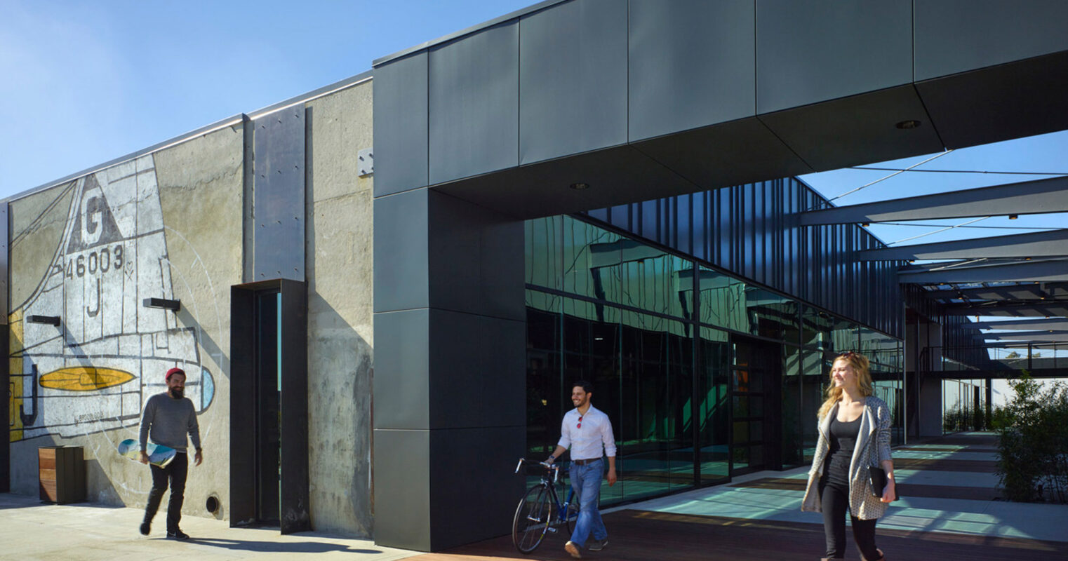 Modern commercial building facade with industrial design elements, featuring weathered concrete panels, expansive glass entrance, and a bold, oversized mural, complemented by an uncluttered wooden walkway and clear blue sky.