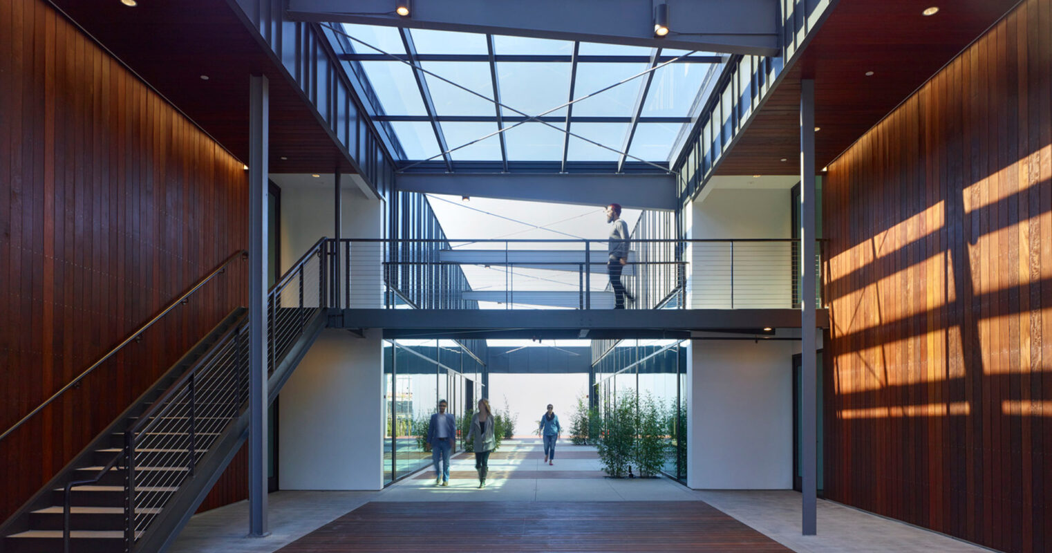 Spacious lobby with floor-to-ceiling wooden panels and a glass roof. A wide staircase to the left leads to a mezzanine where a person walks, while others converse in the sunlit atrium below.