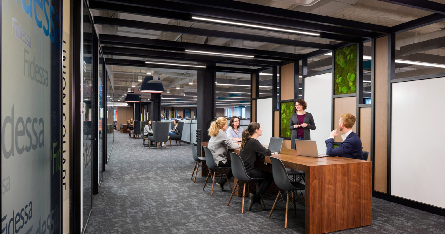 Modern office interior featuring a collaborative workspace with natural wood tables, ergonomic chairs, and privacy booths framed by black metal accents. Ambient lighting and greenery enhance a productive yet comfortable work environment for the group discussion taking place.