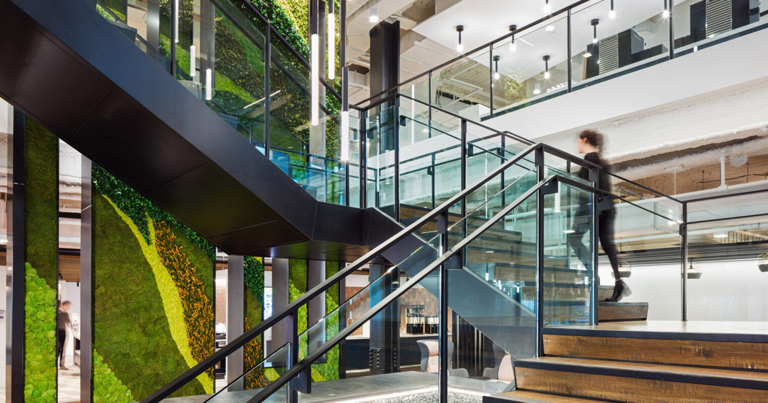 Modern interior with a biophilic design, featuring a spiral staircase with wooden steps, glass balustrades, and living green walls. The space includes concrete ceilings with exposed ductwork, black pendant lighting, and a pebble-filled atrium, emphasizing sustainability and connection to nature.