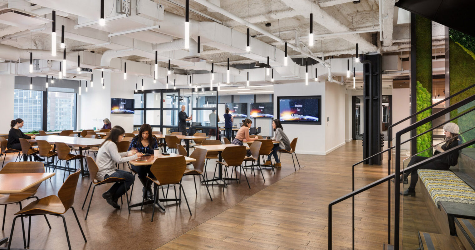 Modern office breakroom featuring an open floor plan with exposed ceilings and industrial lighting. Wooden tables and chairs provide a warm contrast to the concrete textures, while greenery adds a touch of nature to the urban space.