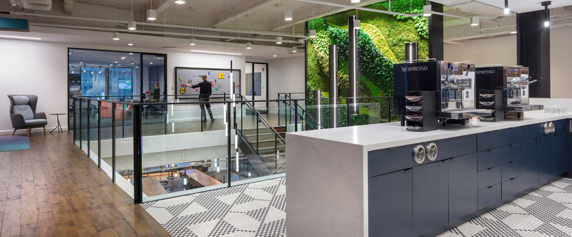 Modern open-plan office space featuring sleek geometric black and white floor tiles, with a full-height living green wall providing a focal point. Glass partitions offer transparency, and exposed beams add an industrial touch, complemented by warm lighting.