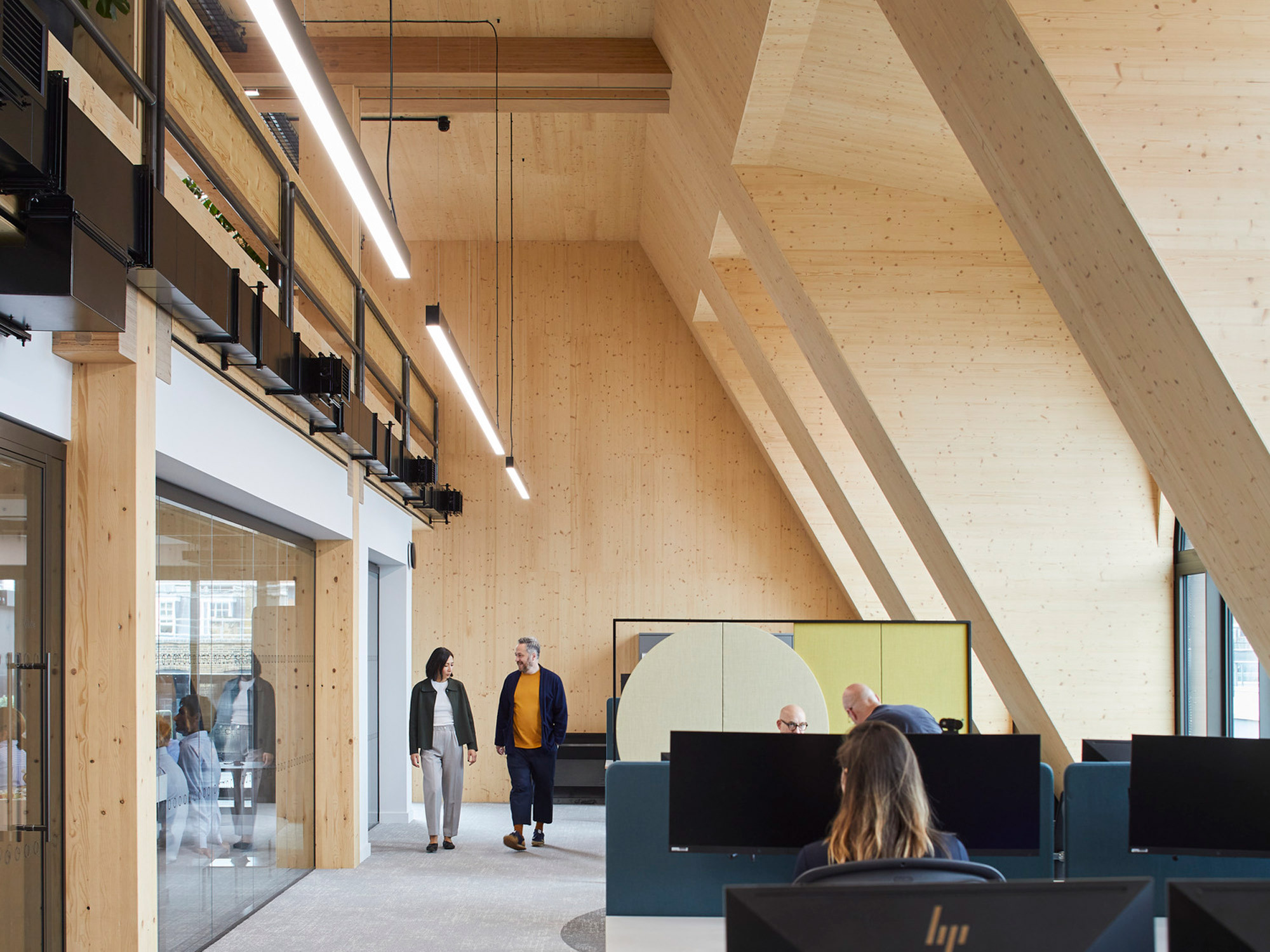 Modern office interior showcasing exposed wooden beams and full-height glazing that floods the multi-tiered workspace with natural light. Open-plan desks are set against a backdrop of warm wooden cladding, enhancing the space's contemporary and inviting atmosphere.