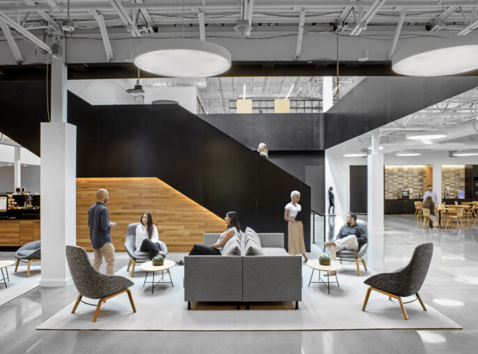 Modern office lounge with monochromatic color scheme, featuring a striking black staircase centerpiece and eclectic seating arrangements with Scandinavian-inspired chairs, promoting collaborative and individual relaxation spaces.