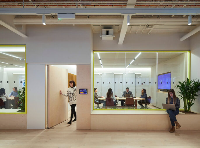Modern office space featuring glass-partitioned meeting rooms with bespoke pendant lighting, exposed ceiling beams, and Ipe wood accents. Strategic use of mirrors expands the visual space, while employees engage in collaborative work highlighted by a warm, neutral color palette.