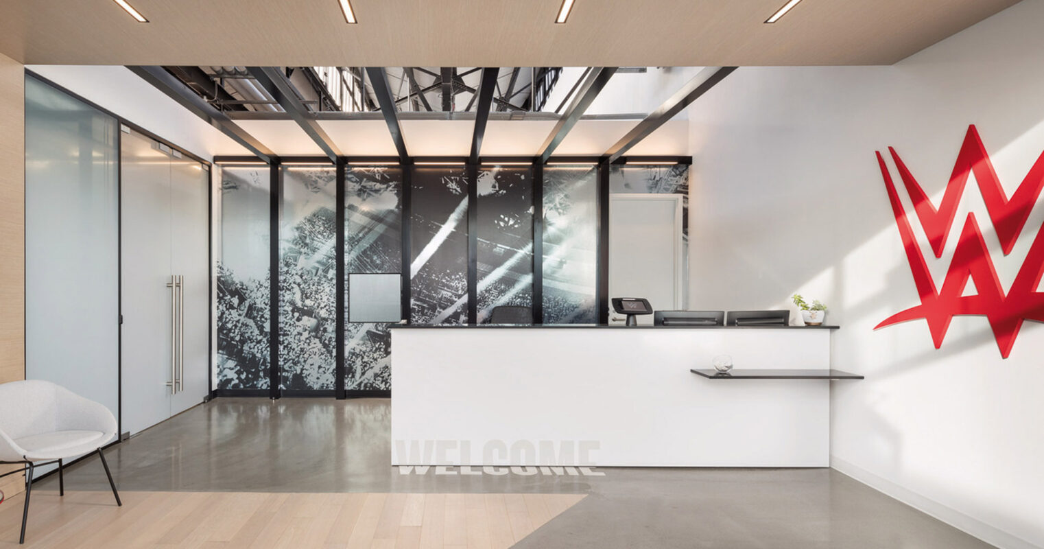 Modern commercial reception area featuring a minimalist design with sleek lines. A white reception desk anchors the space, accompanied by warm wooden flooring and distinctive overhead LED lighting. Full-height glass partitions display monochrome forest graphics, enhancing the open, airy ambiance. A vibrant red wall sculpture adds a pop of color and artistic flair.